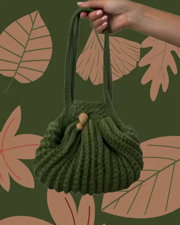 La borsa a sacchetto verde bosco all'uncinetto con due perle, appesa a una mano su un fondo di foglie
